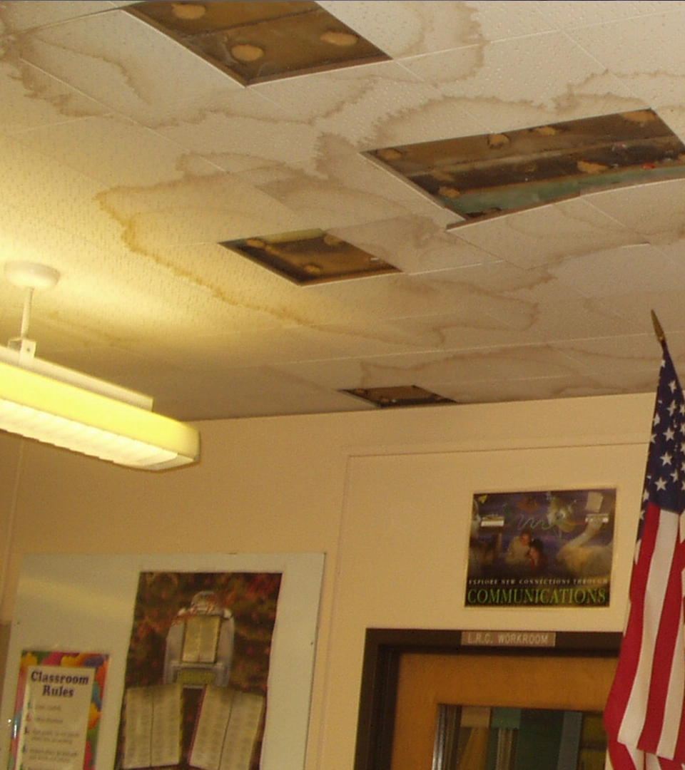 Building condition of indoor ceiling with numerous large water stains and several ceiling tiles removed, exposing brown and moldy areas above; below is part of a classroom wall with an American flag, a door labeled 'LRC Workroom,' and posters including one titled 'Communications.'