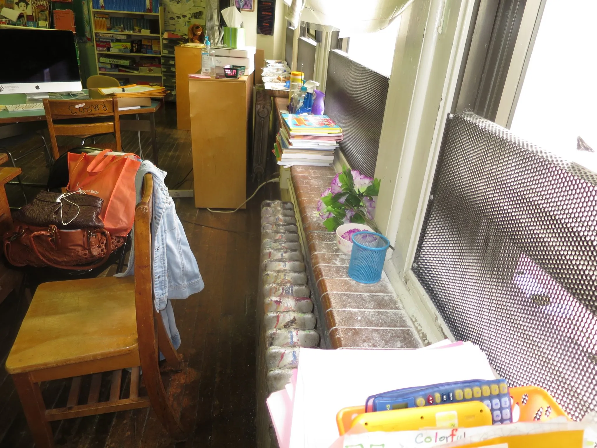 Lead in a classroom interior with wooden chairs, school supplies including calculators and books on a windowsill, a denim jacket on a chair, and a large computer on a desk in the background.